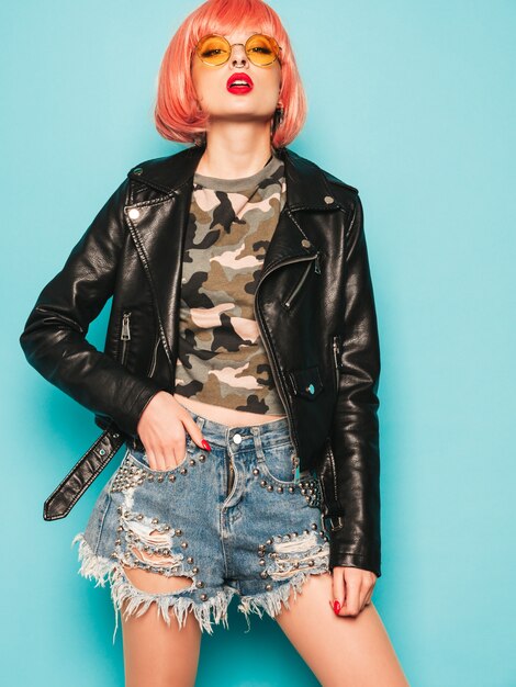 Young beautiful hipster bad girl in black leather jacket and earring in nose.Sexy carefree  woman posing in studio in pink wig near blue wall.Confident model in sunglasses