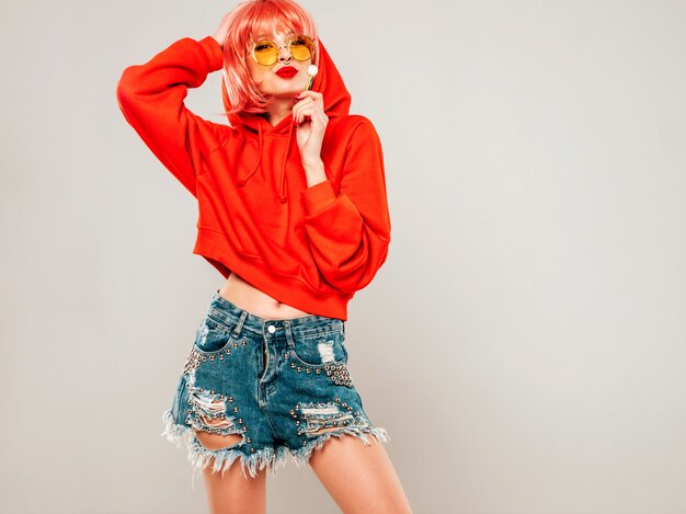 young beautiful hipster bad girl in trendy red summer red hoodie and earring in her nose.sexy carefree woman posing in studio on gray background in wig.hot model licking round sugar candy