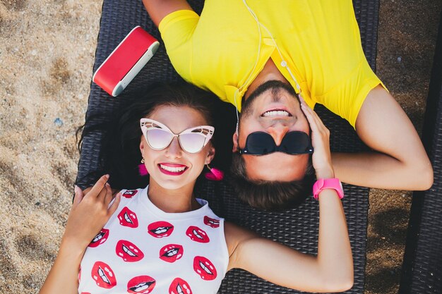 Young beautiful hipster couple in love, lying embracing, listening to music, sunglasses, stylish outfit, summer vacation, having fun, smiling, happy, colorful, view from above