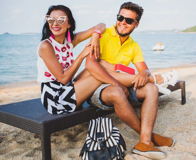 Young beautiful hipster couple in love sitting on beach, listening to music, sunglasses, stylish outfit, summer vacation, having fun, smiling, happy, colorful, positive emotion