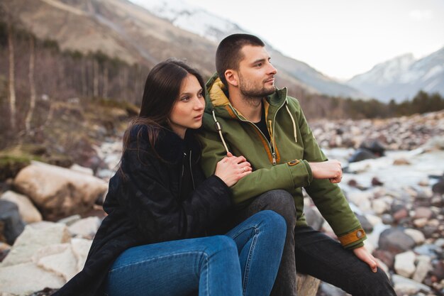 Young beautiful hipster couple in love, sitting by the river, wild nature, winter vacation