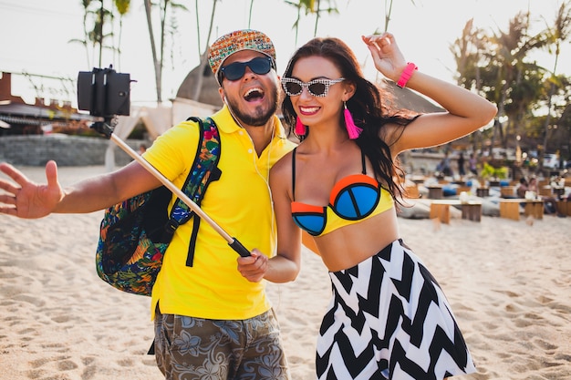 Young beautiful hipster couple in love on tropical beach, taking selfie photo on smartphone, sunglasses, stylish outfit, summer vacation, having fun, smiling, happy, colorful, positive emotion