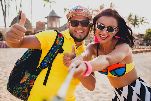 Молодая красивая хипстерская влюбленная пара на тропическом пляже, делающая селфи на смартфоне, солнцезащитные очки, стильный наряд, летние каникулы, весело, улыбаясь, счастливые, красочные, положительные эмоции