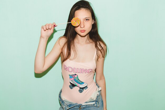 Young beautiful hipster woman posing against blue wall, holding lolly pop candy