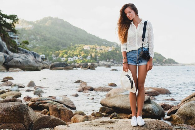 Young beautiful hipster woman on summer vacation in asia, relaxing on tropical beach, digital photo camera, casual boho style, sea landscape , slim tanned body, travel alone