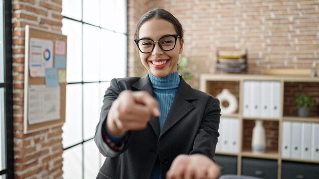 Foto gratuita giovane bellissima donna ispanica lavoratrice d'affari sorridente che indica la telecamera in ufficio