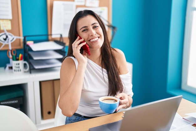 オフィスでコーヒーを飲みながらスマート フォンで話している若い美しいヒスパニック系女性ビジネス ワーカー