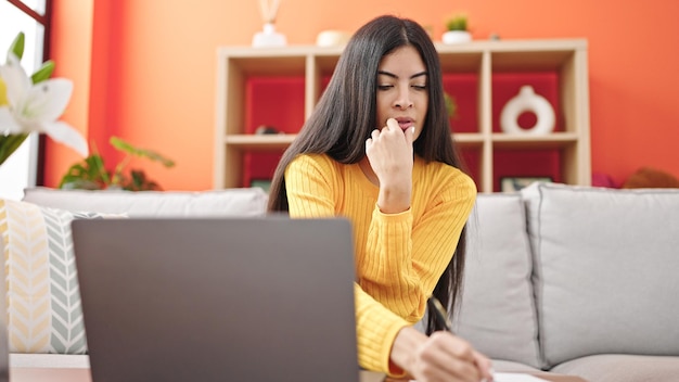 Young beautiful hispanic woman using laptop writing on notebook at home