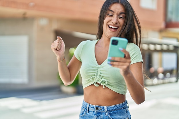 Young beautiful hispanic woman using smartphone with winner expression at street