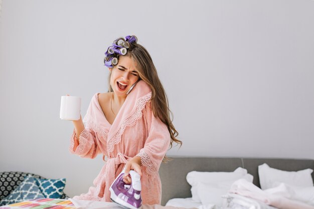 Young beautiful housewife  in pink bathrobe and curler on head holding iron, speaking on phone and holding a cup. She looks astonished.