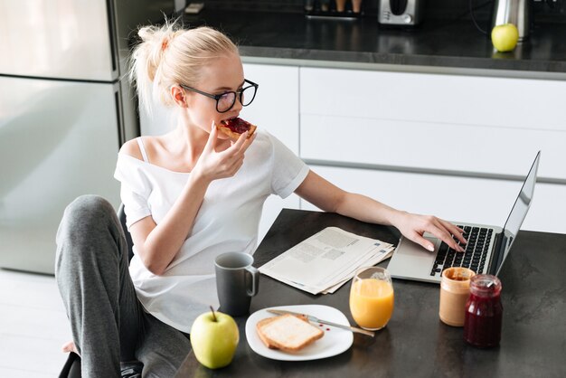 若い美しい女性が朝食をとり、キッチンでラップトップコンピューターを使用して