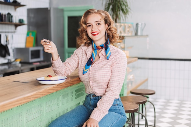 Young beautiful lady in shirt and jeans sitting at the bar counter in cafe and eating cake while happily looking in camera