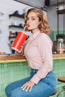 Young beautiful lady in shirt and jeans sitting at the bar counter and drinking soda water while thoughtfully looking in camera in cafe