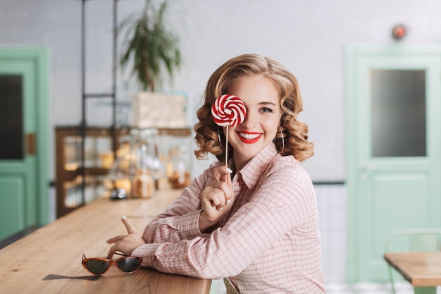 Free photo young beautiful lady sitting at the bar counter and covering her eye with lollipop candy while happily looking in camera in cafe