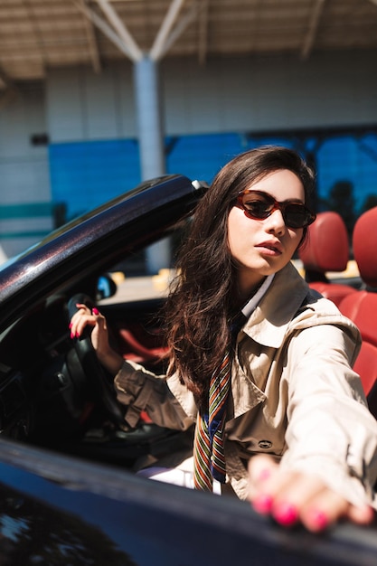 Young beautiful lady in sunglasses and trench coat opening cabriolet car door dreamily looking in camera outdoor