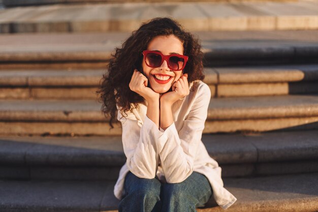 Young beautiful lady with dark curly hair in sunglasses and white jacket sitting on stairs on street and happily looking in camera