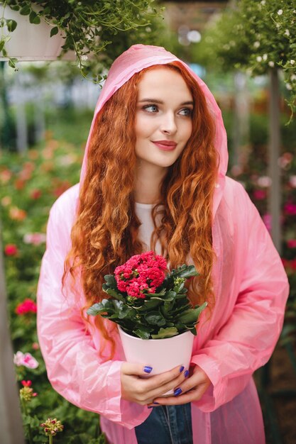 Young beautiful lady with redhead curly hair standing in pink raincoat and holding flower in pot in hands while dreamily looking aside in greenhouse