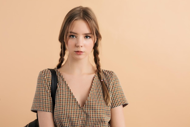 Young beautiful lady with two braids in tweed dress with black backpack on shoulder thoughtfully looking in camera over beige background isolated