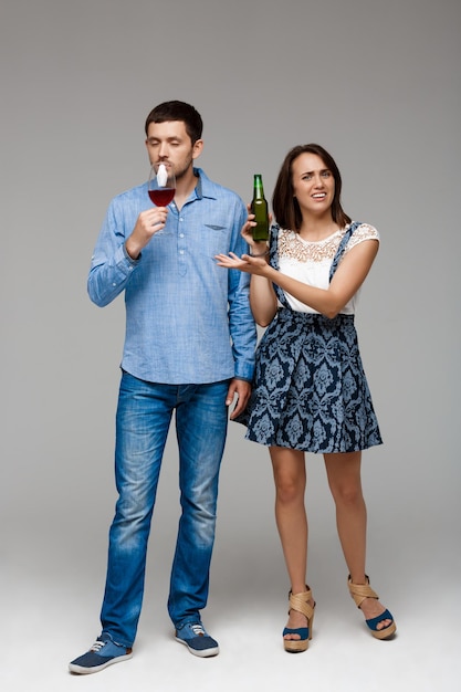 Young beautiful married couple drinking wine and beer over grey wall