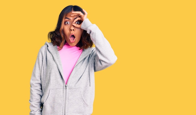 Young beautiful mixed race woman wearing casual sporty clothes doing ok gesture shocked with surprised face, eye looking through fingers. unbelieving expression.