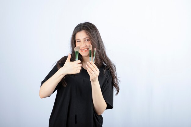 young beautiful model with makeup with brushes posing on white wall. 