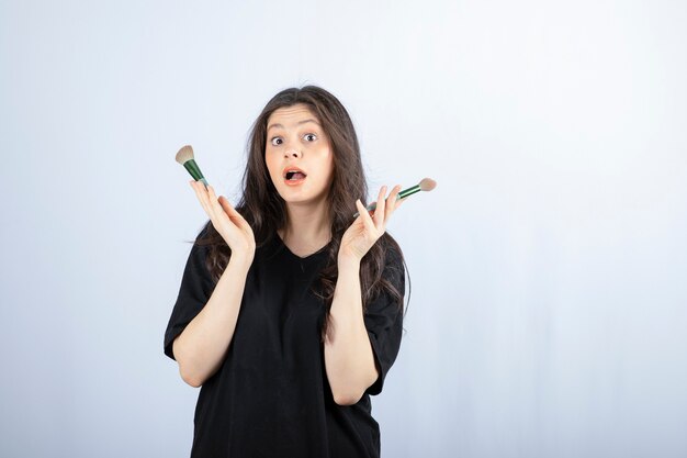 young beautiful model with makeup with brushes posing on white wall. 