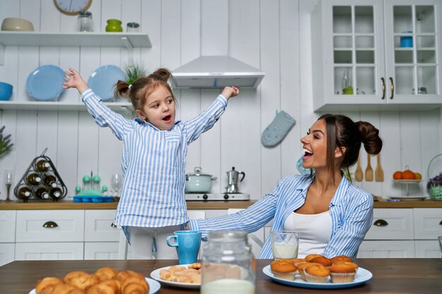 若い美しい母親と彼女の小さな娘は、自宅で朝食時にキッチンで遊んでいます。