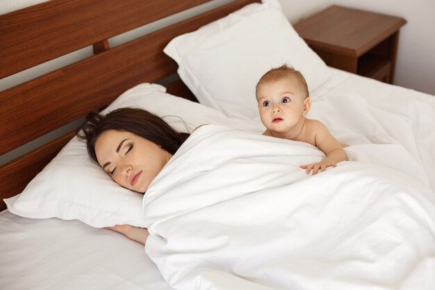 Молодая красивая мать и ее спать новорожденного младенца лежа в кровати рано утром.
