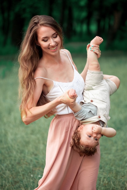 Young beautiful mother hugging her little toddler son against green grass. Happy woman with her baby boy on a summer sunny day. Family walking on the meadow.