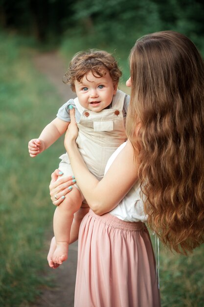 Young beautiful mother hugging her little toddler son against green grass. Happy woman with her baby boy on a summer sunny day. Family walking on the meadow.