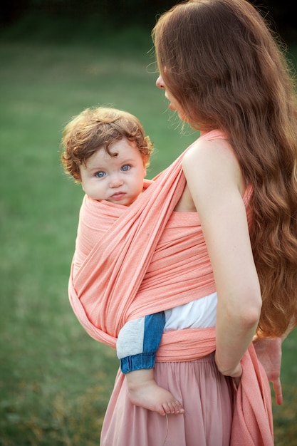 Young beautiful mother hugging her little toddler son against green grass. Happy woman with her baby boy on a summer sunny day. Family walking on the meadow.