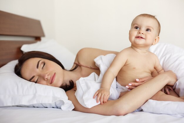 Young beautiful mother with closed eyes and her newborn baby lying in bed early morning.