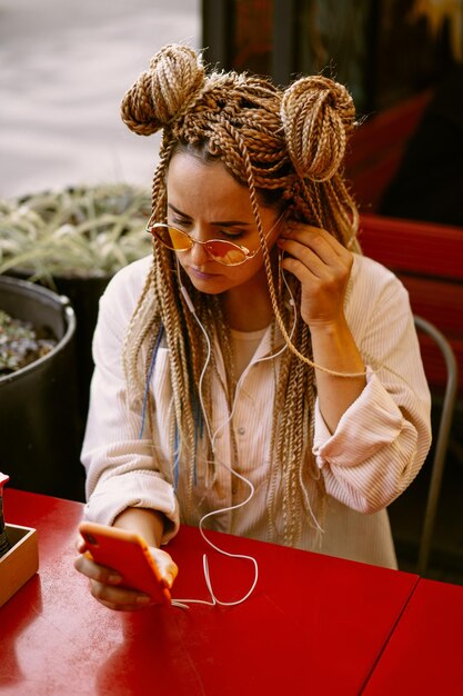 Young beautiful multiracial woman with blonde afro hairstyle braids zizi on the street cafe with headphones. yellow sunglasses, bright makeup, hippie style. video call, work outside the home