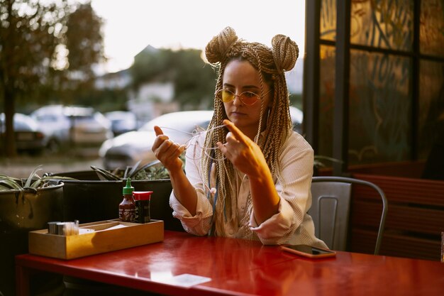 Young beautiful multiracial woman with blonde afro hairstyle braids zizi on the street cafe with headphones. yellow sunglasses, bright makeup, hippie style