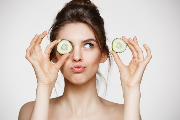 Young beautiful naked girl smiling hiding eye behind cucumber slice over white background. Beauty spa and cosmetology concept.