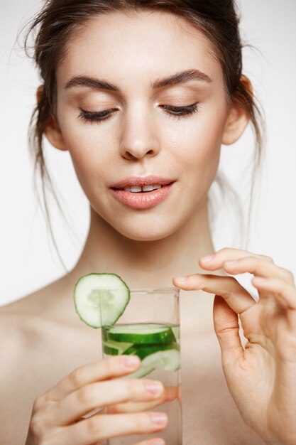 Young beautiful naked girl with perfect clean skin smiling holding glass of water with cucumber slices over white background. Facial treatment.