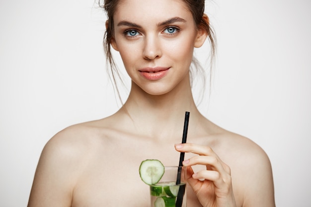 Young beautiful naked girl with perfect clean skin smiling looking at camera holding glass of water with cucumber slices over white background. Facial treatment.