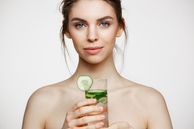 Young beautiful naked girl with perfect clean skin smiling looking at camera holding glass of water with cucumber slices over white background. Facial treatment.