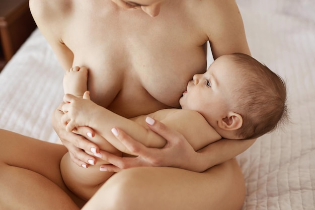Young beautiful naked mom breastfeeding hugging her newborn baby smiling sitting on bed at home. Copy space.