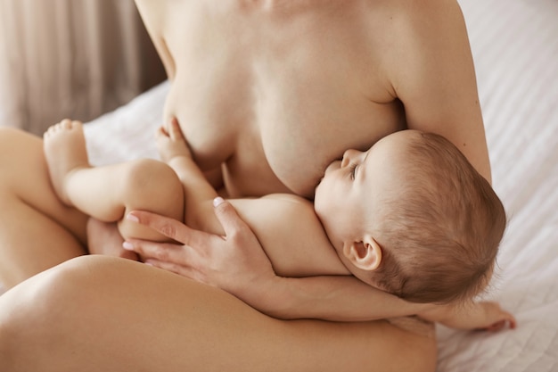 Молодая красивая голая мама кормит грудью, обнимая своего новорожденного ребенка, улыбаясь сидя на кровати у себя дома