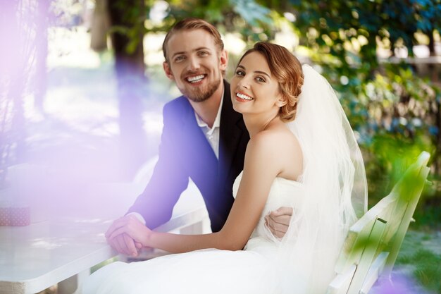 Young beautiful newlyweds smiling, embracing, sitting in cafe outdoors.