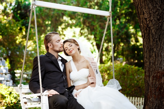 Young beautiful newlyweds smiling, kissing, sitting on swing in park.
