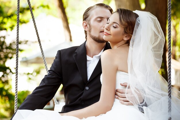 Young beautiful newlyweds smiling, kissing, sitting on swing in park.