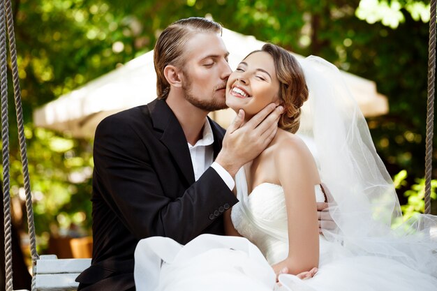 Young beautiful newlyweds smiling, kissing, sitting on swing in park.