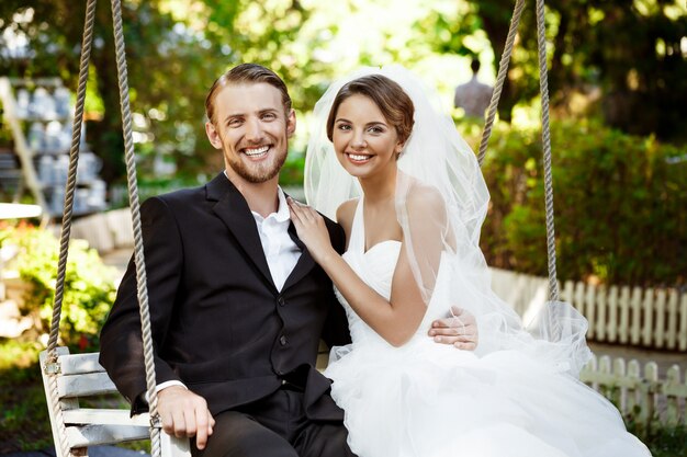 Young beautiful newlyweds smiling, laughing, sitting on swing in park.