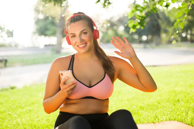 ピンクのスポーティなトップと赤いヘッドフォンでレギンスの若い美しいプラスサイズの女性は、カメラを見て、都市公園で携帯電話を手に手を振っています