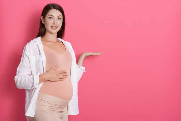 コピースペースを示す手の開いた手のひらで脇を指しているピンク色の空間を分離した赤ちゃんを期待して美しい妊娠中の少女は、広告を提示し、カメラに微笑んでいます。