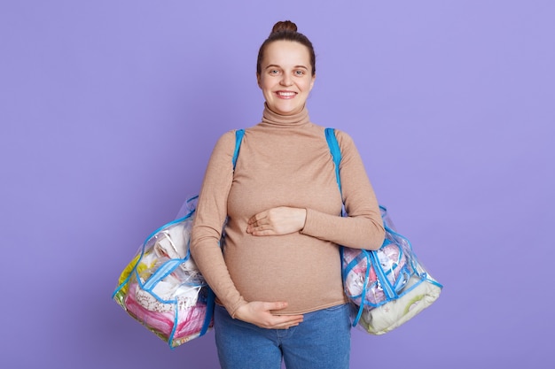 Young beautiful pregnant woman standing isolated over lilac wall,, touching her belly, holding bags with stuff for maternity house.