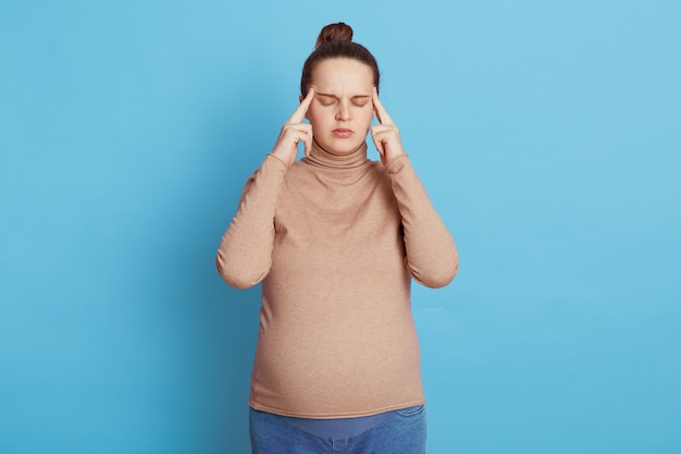 Young beautiful pregnant woman with closed eyes expecting baby, keeping fingers on temples, thinks about something important, poses isolated on blue wall, being confused and annoyed.