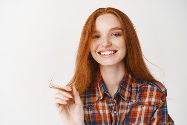 Giovane bella donna rossa che sorride con i denti bianchi perfetti alla telecamera toccando una ciocca di capelli allo zenzero e guardando felice in piedi su sfondo bianco